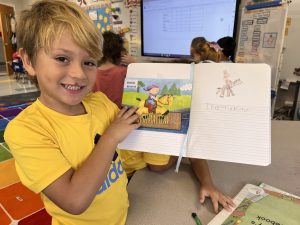 Kindergarten student smiles while holding up his book and writing notebook