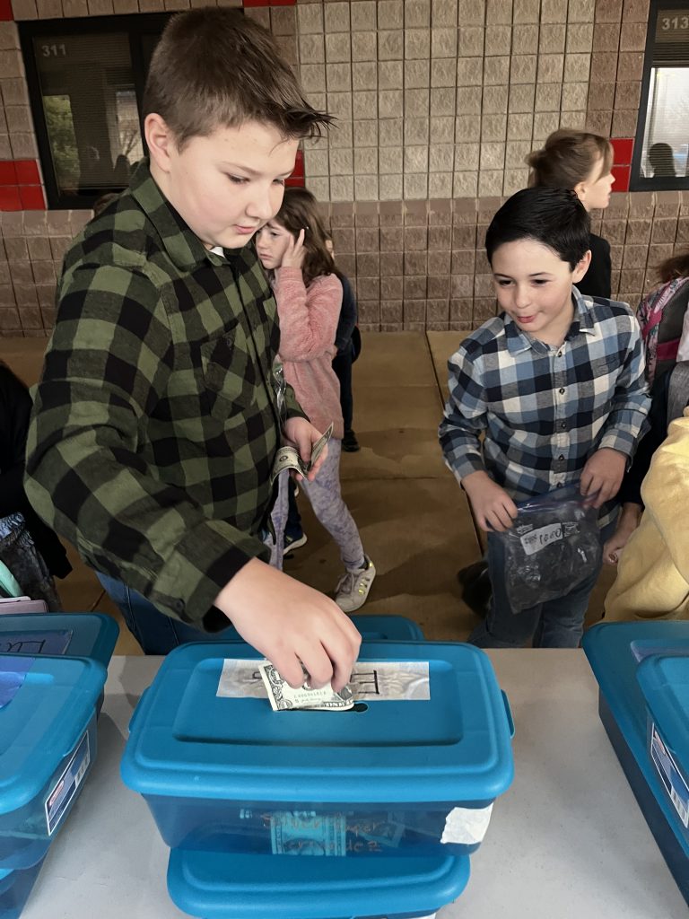 Fifth grade student putting a dollar bill into a fourth grade class bucket which will subtraction from their overall penny count.