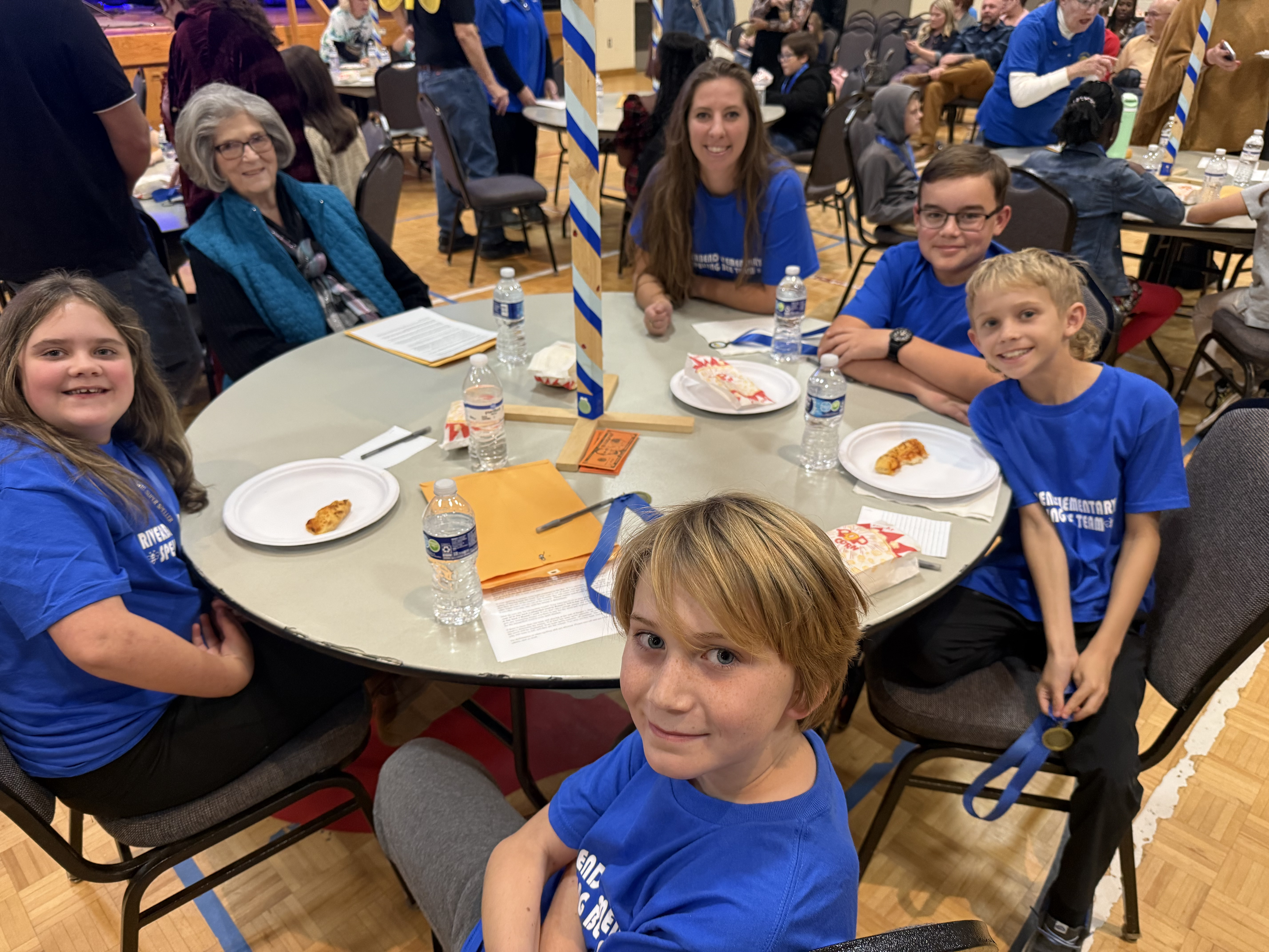 Group of 4 students, 1 teacher, and 1 volunteer sitting around a table getting ready for the Spelling Bee to strart.
