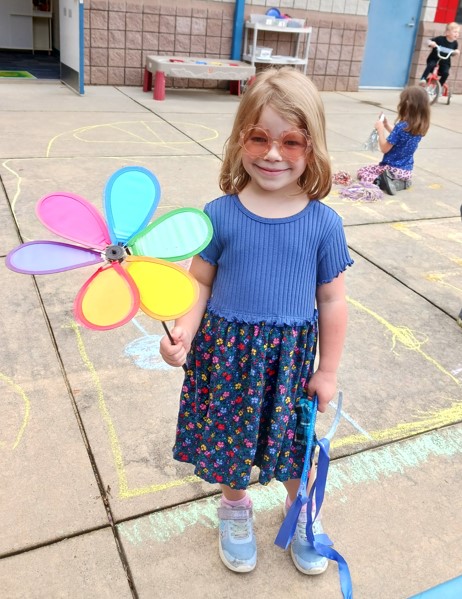 Young girl with blue flower dress and pink heart sunglasses standing outside on a playground holding a rainbow colored pinwheel flower and streamers.  In the background are other children playing.  