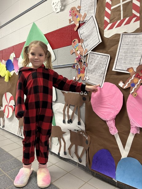 Young girl with blonde pigtales wearing plaid pajamas and pink slippers pointing to her writing on a gingerbread house in the hallway of a school. 
