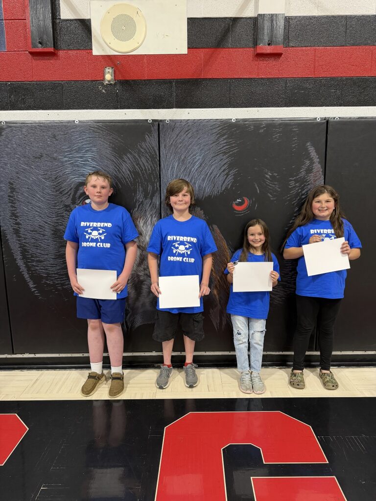 Four students, two boys and two girls, standing in front of a black gym mat hanging on the wall with a black bear sketch on it.  Floor and wall are also painting black and read. Student's t-shirts read RIVERBEND DRONE CLUB.  Each student is holding their certificate and drone legend wings pin. 