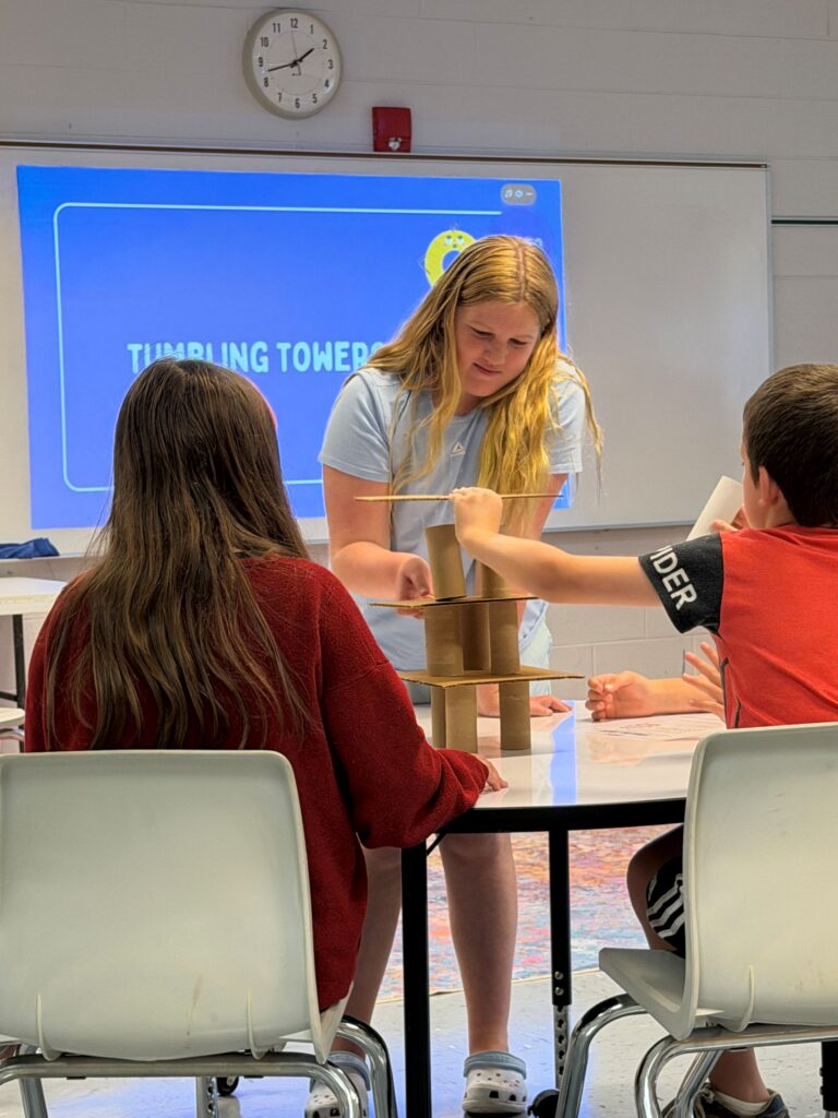 Students work in a small group to complete an engineering task.  Two students are seated at a round table helping another student who is  standing move a paper towel roll and a piece of cardboard to create a tower and keep it from tumbling. 