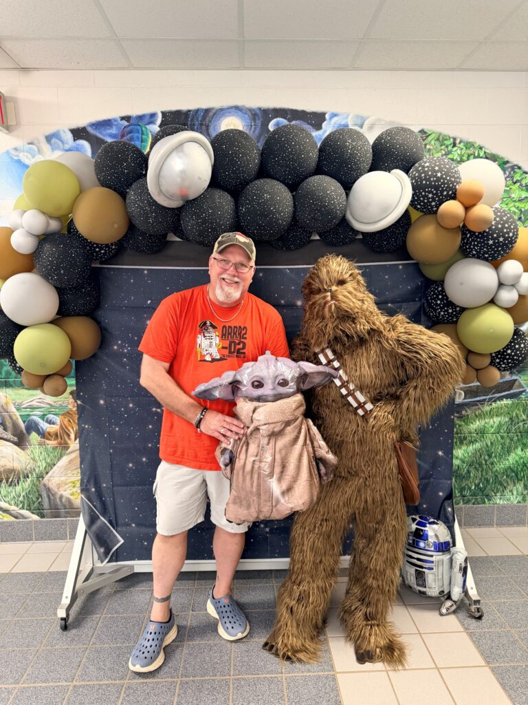 Male Teacher standing in front of a space themed backdrop.  Holding a balloon alien character.  Beside him is a Chewbacca.  