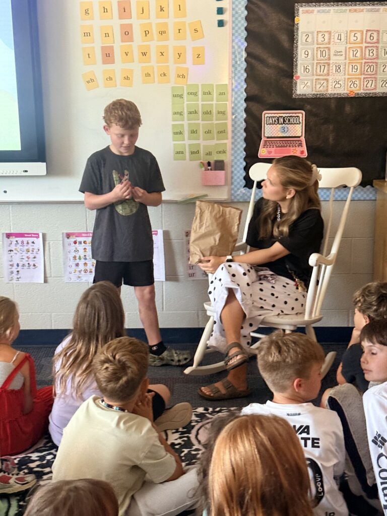 A student is in front of his classmates sharing items from a brown paper bag to explain about himself.  A teacher sits in the rocking chair beside him listening to him share his "Me Bag." Classmates sit on the floor facing the student and listen.