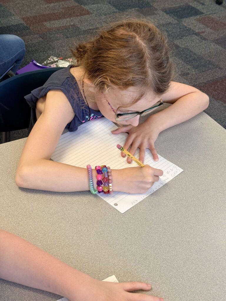 A student is sitting at a table writing on lined paper with a pencil. 