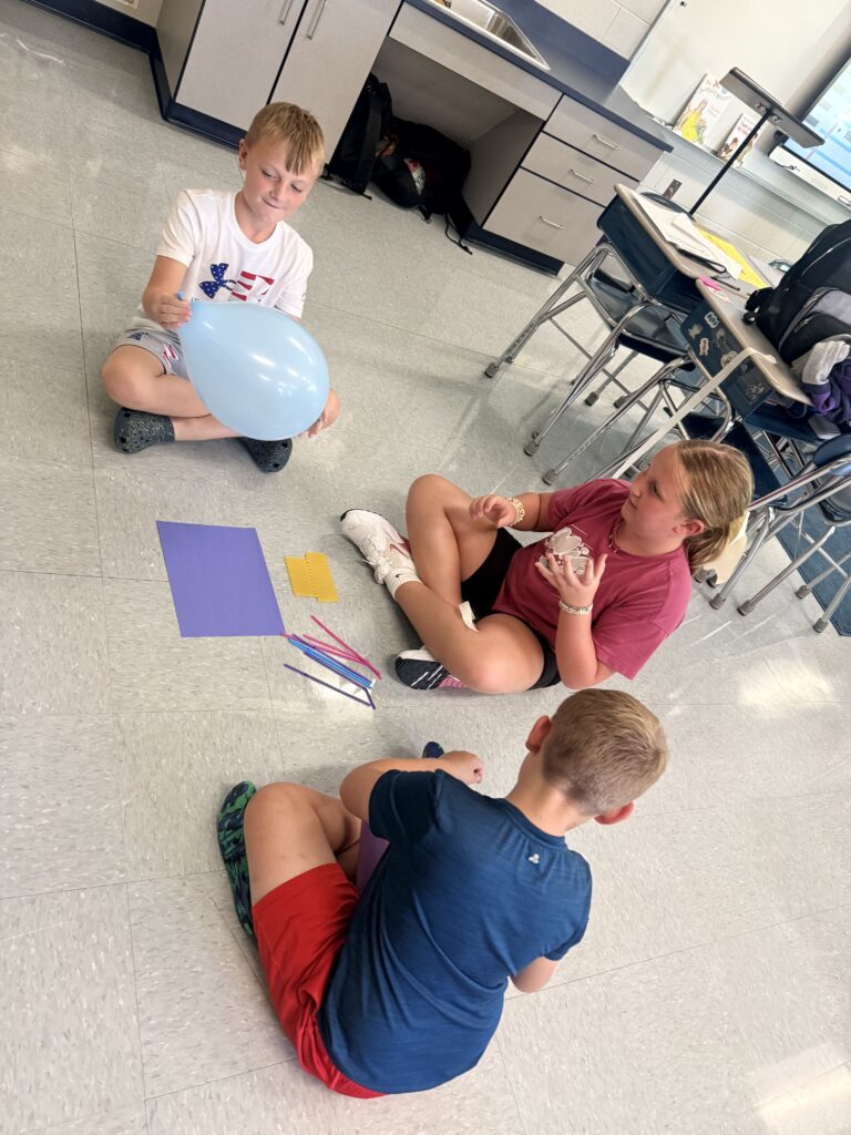 Three students sit on the floor of their classroom.  One student is holding a blue balloon.  Another student has tape in her hands.  There are pieces of colorful paper on the floor.