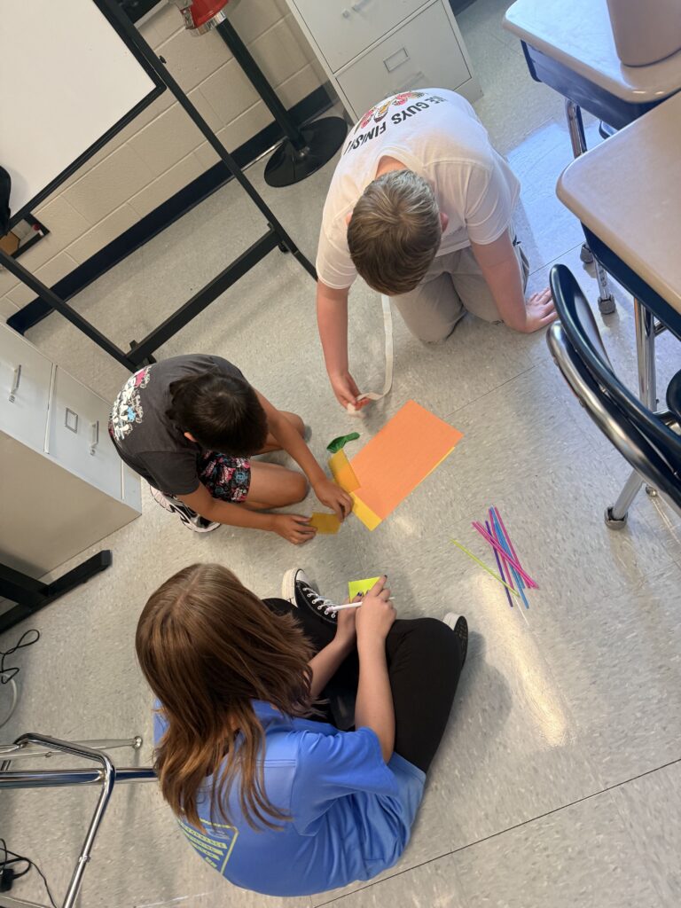 Three students are sitting on the floor in their classroom.  They have brightly colored pieces of paper in front of them.  They are working together to solve the challenge. 