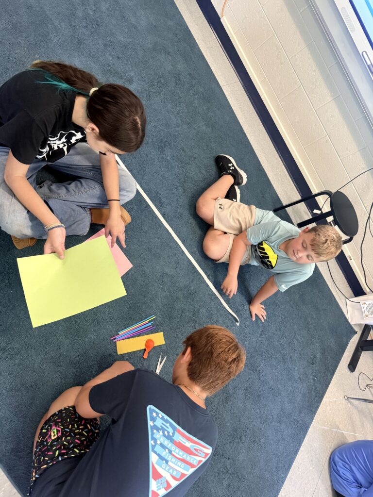 Three students are sitting on the carpeted floor in their classroom.  They have brightly colored pieces of paper in front of them.  They are working together to solve the challenge. 