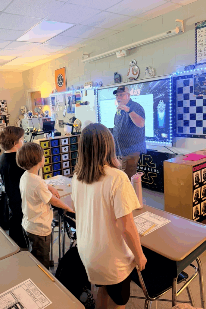 A teacher doing a fun beginning of the school activity with his students.  He is standing at front of the classroom holding the letter R.  Students are standing facing him.  